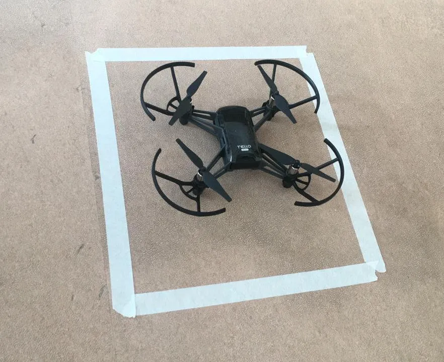 Un dron Tello negro, con protectores circulares en sus cuatro hélices, apoyado en el piso dentro de un cuadrado delimitado con cinta de papel.
