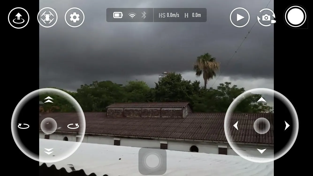 Vista del modo manual de la app Tello: la imagen que transmite la cámara del dron (un cielo nublado sobre construcciones y árboles) con los dos joysticks circulares superpuestos, indicadores de batería, WiFi y altura arriba, y los botones de foto y grabación a la derecha.